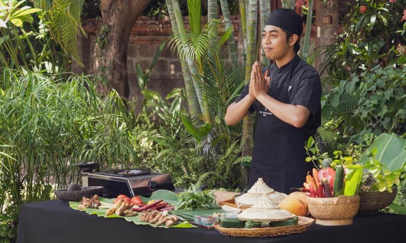 A Balinese chef stands with hands pressed in greeting beside a table of fresh ingredients during an outdoor cooking class. 