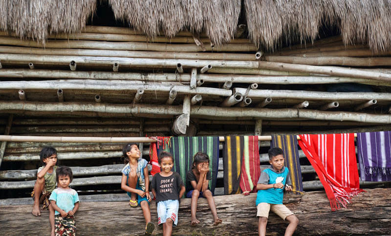 Local children sit by a bamboo hut in a traditional village in Flores, surrounded by colourful handwoven textiles hanging behind them. 
