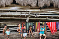Local children sit by a bamboo hut in a traditional village in Flores, surrounded by colourful handwoven textiles hanging behind them. 