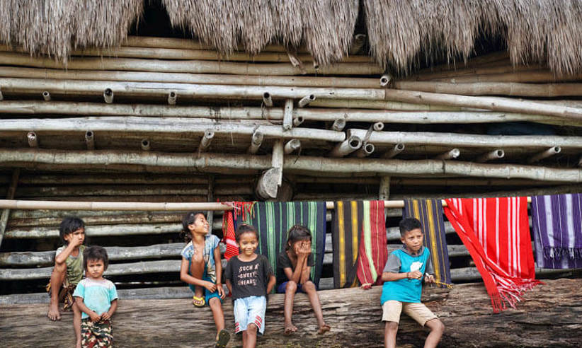 Local children sit by a bamboo hut in a traditional village in Flores, surrounded by colourful handwoven textiles hanging behind them. 