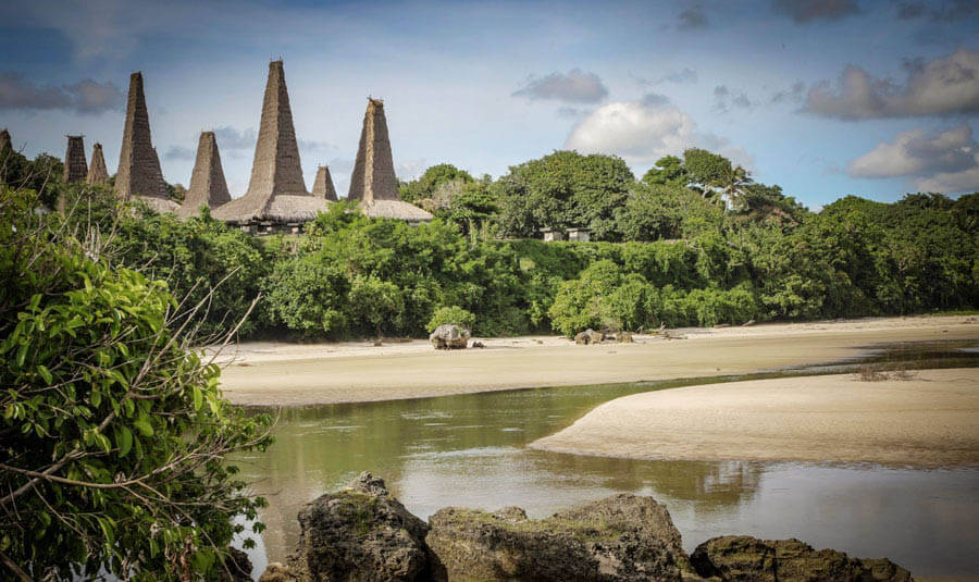 Traditional conical-roof houses of Ratenggaro village in Sumba, Indonesia, seen across a quiet inlet with lush tropical trees. 