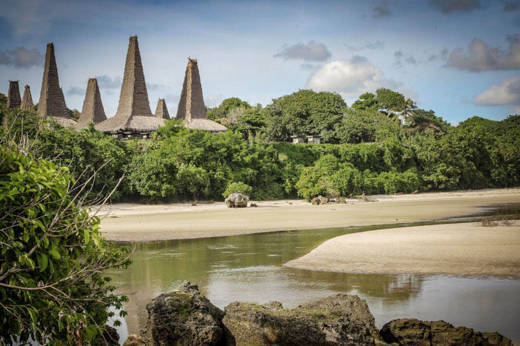 Traditional conical-roof houses of Ratenggaro village in Sumba, Indonesia, seen across a quiet inlet with lush tropical trees. 
