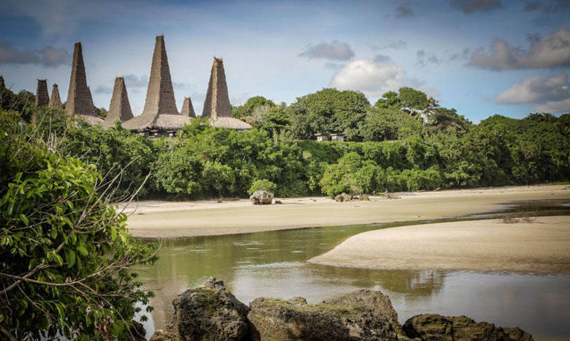 Traditional conical-roof houses of Ratenggaro village in Sumba, Indonesia, seen across a quiet inlet with lush tropical trees. 