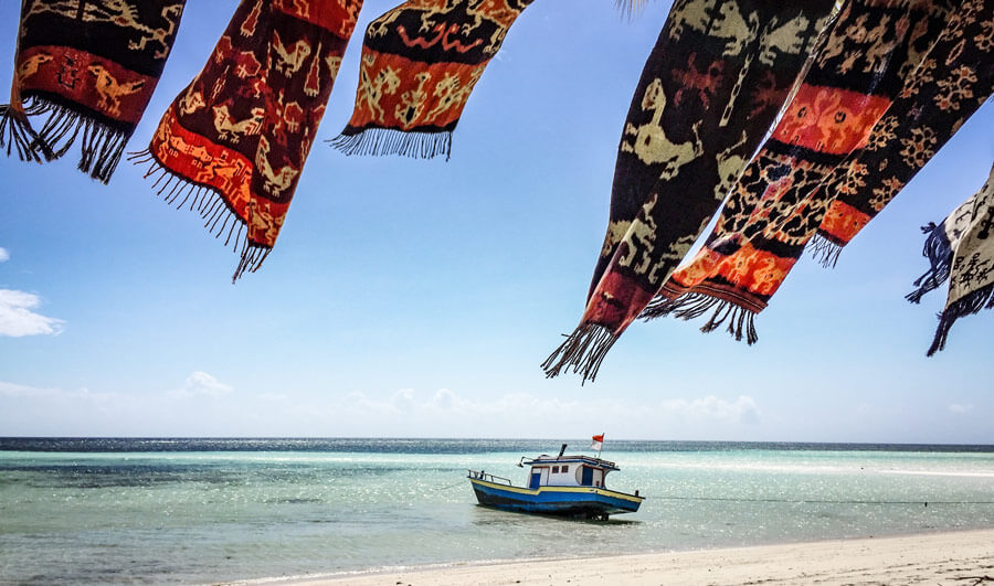Colourful woven textiles flutter over Walakiri Beach, Sumba, with a small boat anchored on calm turquoise waters under clear skies. 