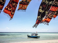 Colourful woven textiles flutter over Walakiri Beach, Sumba, with a small boat anchored on calm turquoise waters under clear skies. 