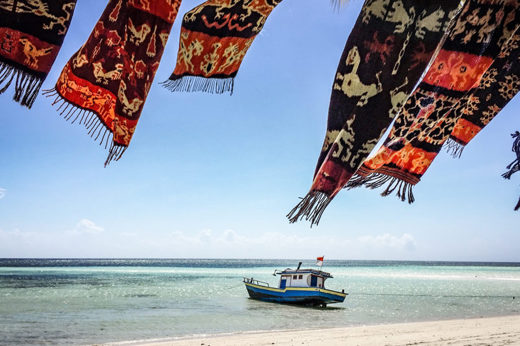 Colourful woven textiles flutter over Walakiri Beach, Sumba, with a small boat anchored on calm turquoise waters under clear skies. 