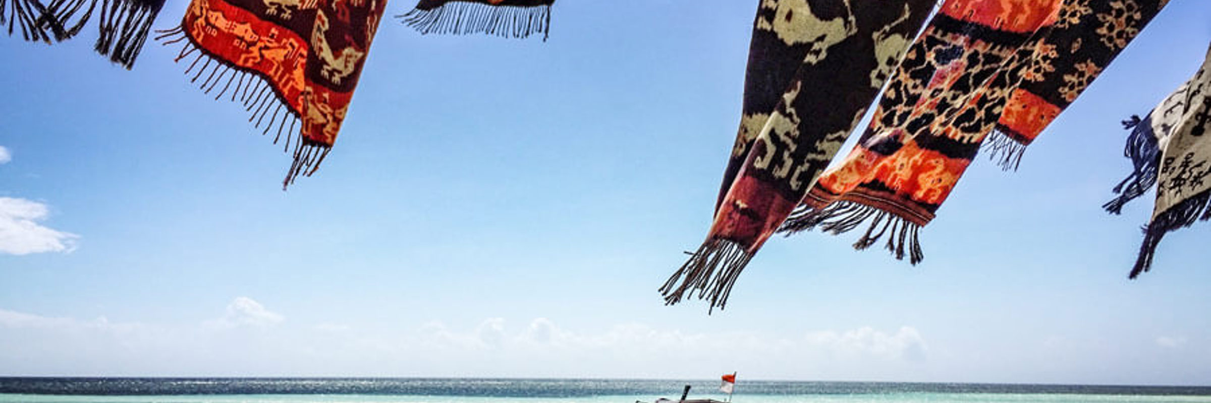 Colourful woven textiles flutter over Walakiri Beach, Sumba, with a small boat anchored on calm turquoise waters under clear skies. 