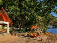 Secluded beachfront bungalow in Indonesia with shaded veranda, loungers, and views of the ocean framed by lush greenery. 