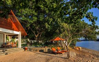 Secluded beachfront bungalow in Indonesia with shaded veranda, loungers, and views of the ocean framed by lush greenery. 