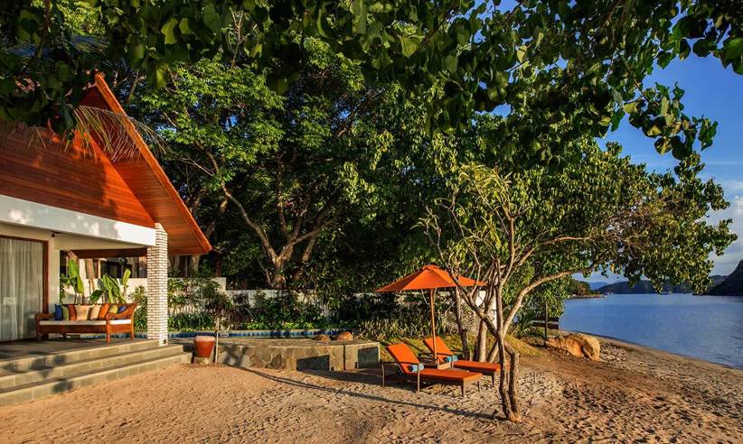 Secluded beachfront bungalow in Indonesia with shaded veranda, loungers, and views of the ocean framed by lush greenery. 