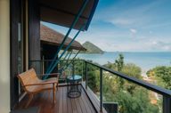 Private balcony suite in Flores, Indonesia with wooden seating, ocean views, and tropical greenery under clear blue skies. 