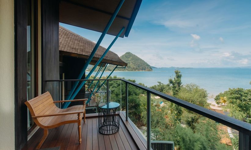 Private balcony suite in Flores, Indonesia with wooden seating, ocean views, and tropical greenery under clear blue skies. 
