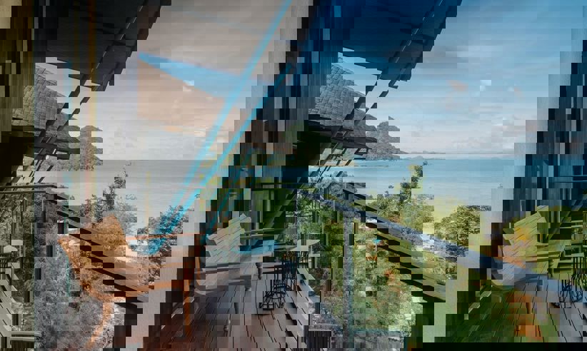 Private balcony suite in Flores, Indonesia with wooden seating, ocean views, and tropical greenery under clear blue skies. 