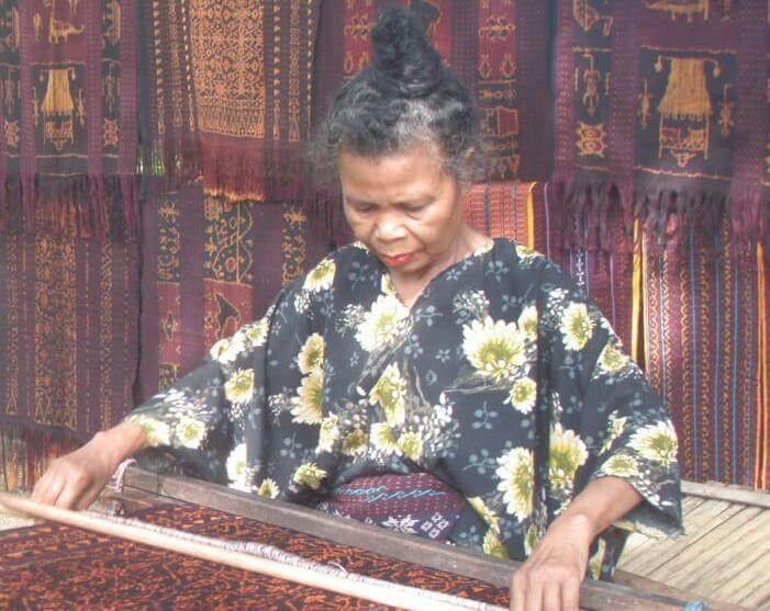 Indonesian woman weaving traditional textiles by hand in Flores, surrounded by rich patterned fabrics in a cultural setting. 