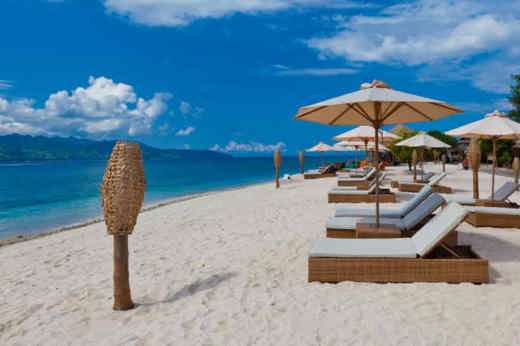 Beachfront sunbeds with umbrellas on the white sands of Gili Trawangan, Lombok, facing turquoise ocean under blue sky. 