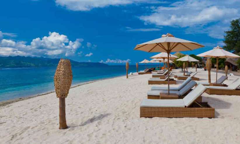 Beachfront sunbeds with umbrellas on the white sands of Gili Trawangan, Lombok, facing turquoise ocean under blue sky. 