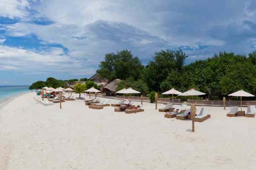 Luxury beach resort on Gili Trawangan with white sand, sun loungers, thatched umbrellas, and lush tropical trees lining the shore. 