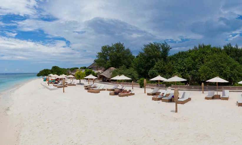 Luxury beach resort on Gili Trawangan with white sand, sun loungers, thatched umbrellas, and lush tropical trees lining the shore. 
