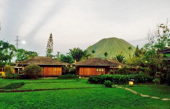 Wooden bungalows sit in a lush green valley in Indonesia, with a misty mountain backdrop and wide open grassy lawn. 