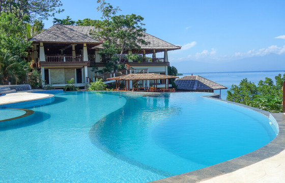 An infinity pool overlooks the turquoise sea beside a luxury villa on a sunny clifftop in Bali, Indonesia. 