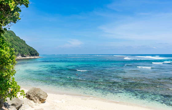 Crystal-clear turquoise water and soft white sand at Sanur Beach, Bali, framed by lush greenery and coral rocks. 