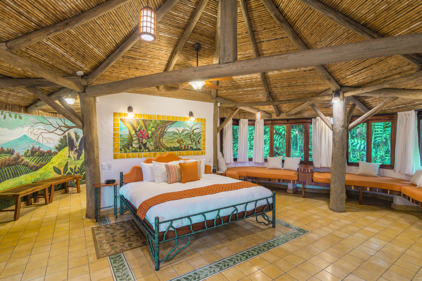 Spacious rustic bedroom at El Ranchito Suite featuring a thatched roof, vibrant artwork, and large windows overlooking the forest. 