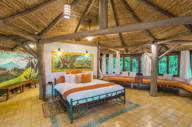 Spacious rustic bedroom at El Ranchito Suite featuring a thatched roof, vibrant artwork, and large windows overlooking the forest. 