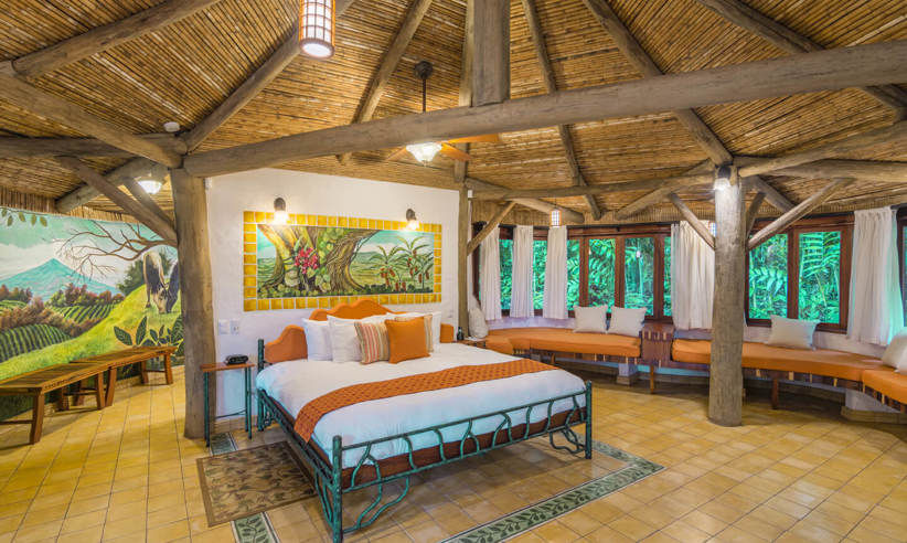 Spacious rustic bedroom at El Ranchito Suite featuring a thatched roof, vibrant artwork, and large windows overlooking the forest. 