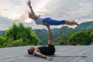A couple performs partner yoga on a wooden deck, framed by lush rainforest and a tranquil Costa Rican sunset. 
