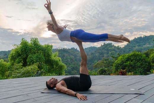 A couple performs partner yoga on a wooden deck, framed by lush rainforest and a tranquil Costa Rican sunset. 