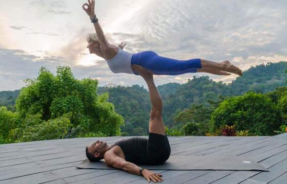 A couple performs partner yoga on a wooden deck, framed by lush rainforest and a tranquil Costa Rican sunset. 