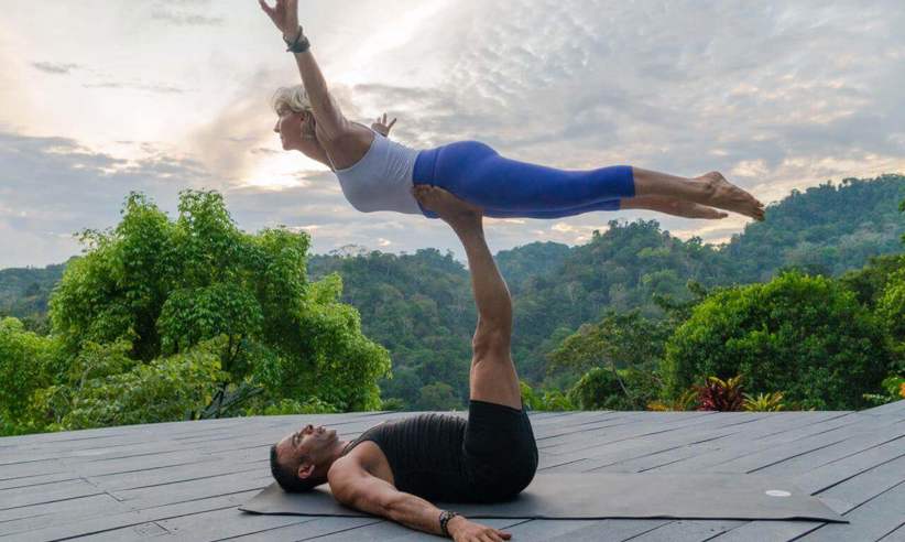 A couple performs partner yoga on a wooden deck, framed by lush rainforest and a tranquil Costa Rican sunset. 