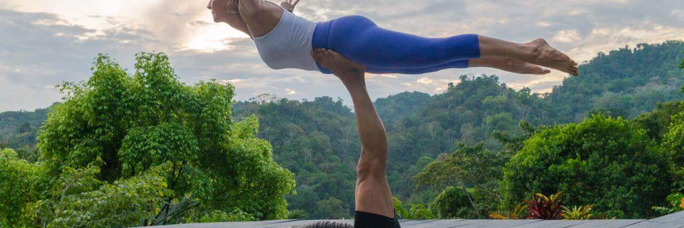 A couple performs partner yoga on a wooden deck, framed by lush rainforest and a tranquil Costa Rican sunset. 