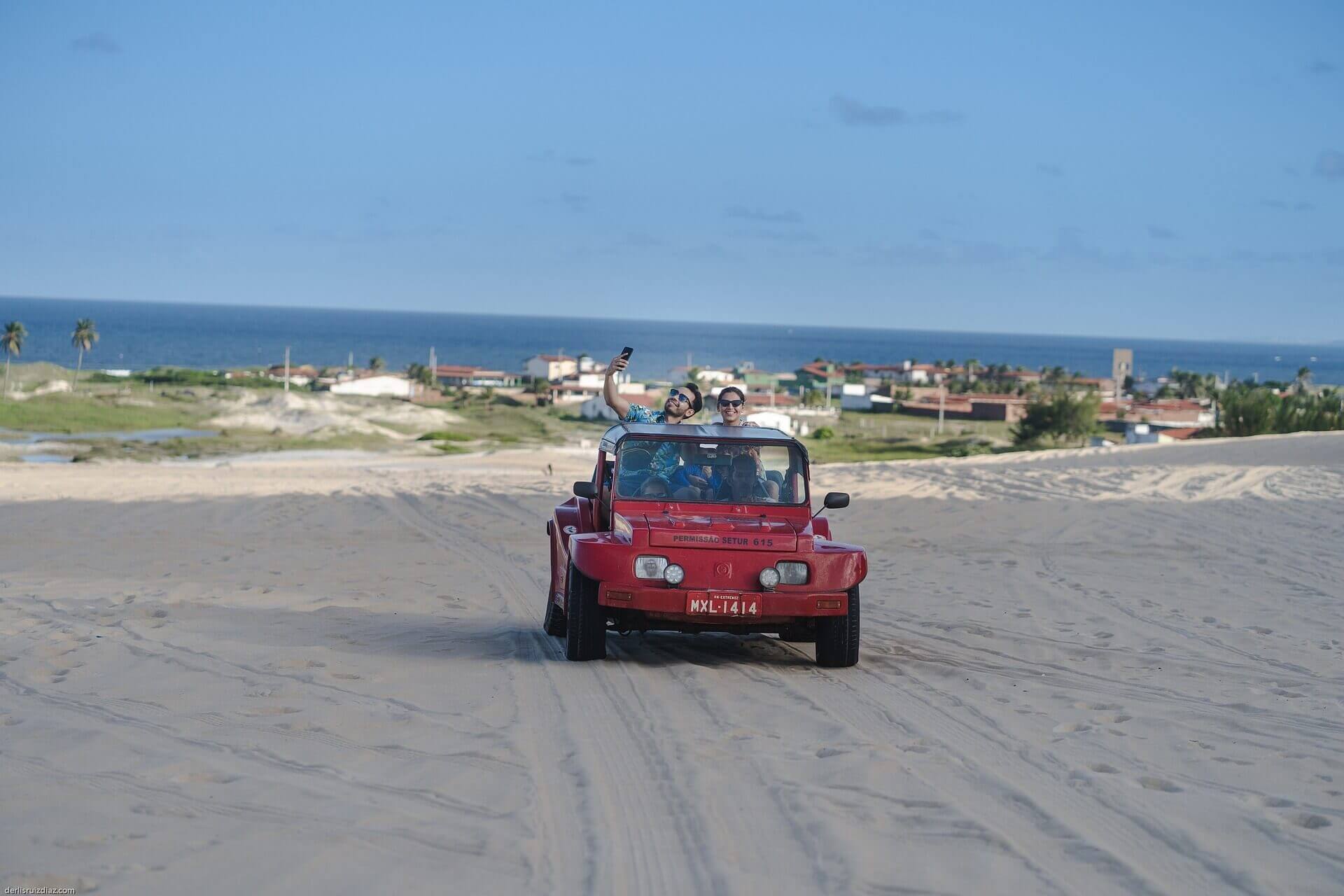 Red beach buggy driving  across sand dunes in Jericoacoara with ocean and coastal town in the background. 
