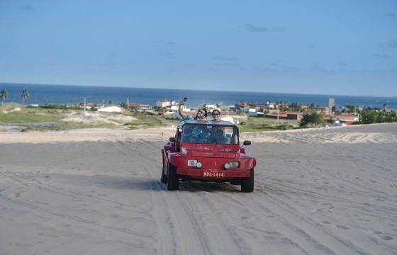 Red beach buggy driving  across sand dunes in Jericoacoara with ocean and coastal town in the background. 