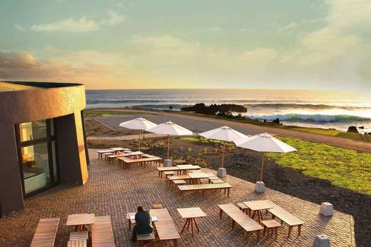 Outdoor terrace at Nayara Easter Island overlooks the ocean at sunset with tables, parasols, and scenic views. 