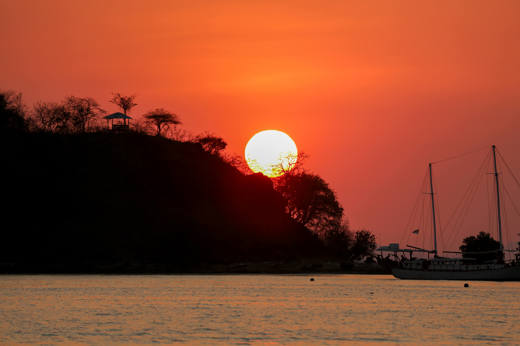 Fiery sunset over Flores Island with Silhouetted trees and a sailing boat on calm waters, casting reflections in Indonesia. 