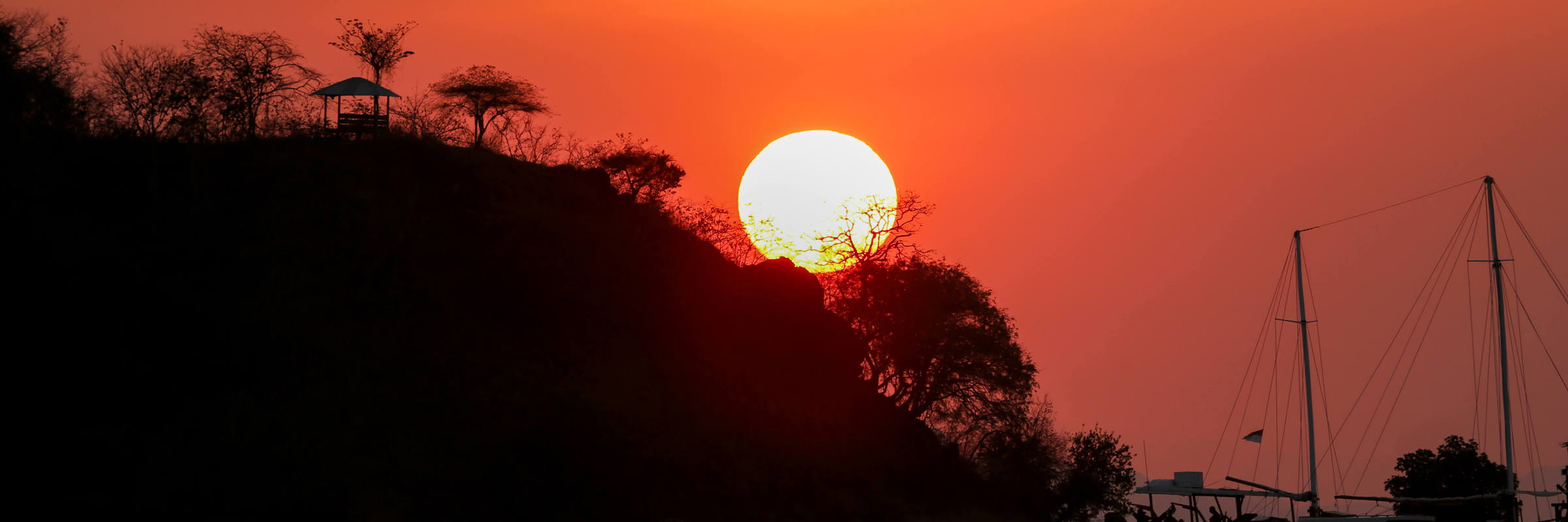 Fiery sunset over Flores Island with Silhouetted trees and a sailing boat on calm waters, casting reflections in Indonesia. 