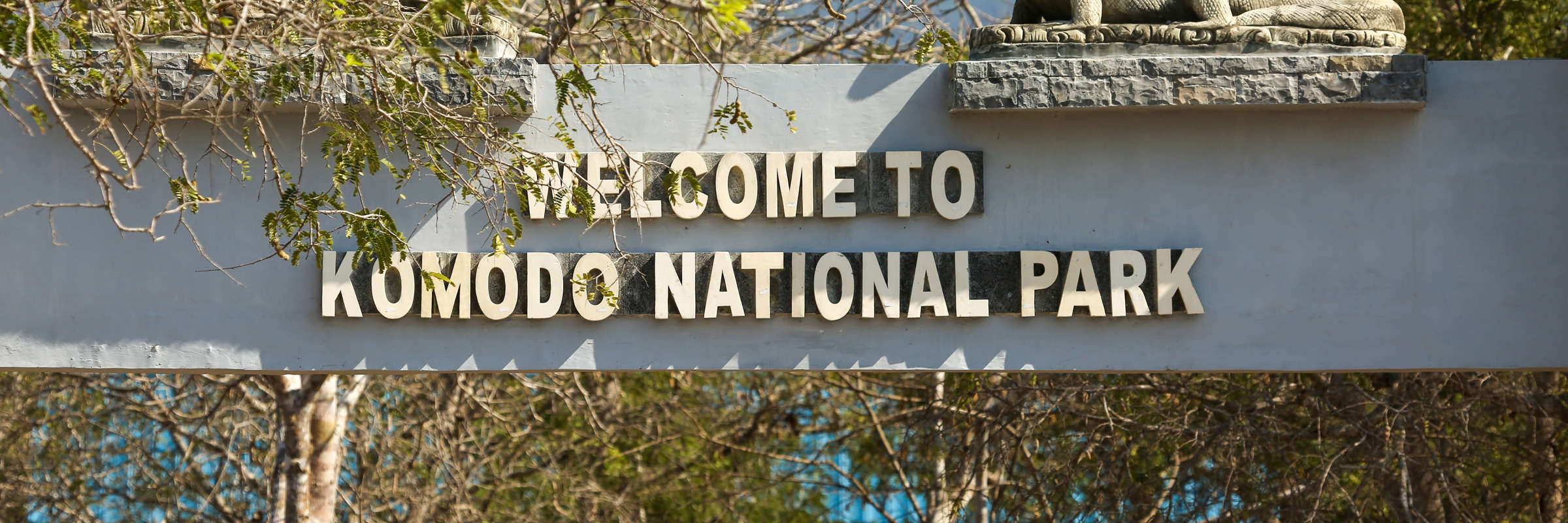 Entrance sign to Komodo National Park in Indonesia, featuring a carved Komodo dragon statue and shaded by forest trees. 