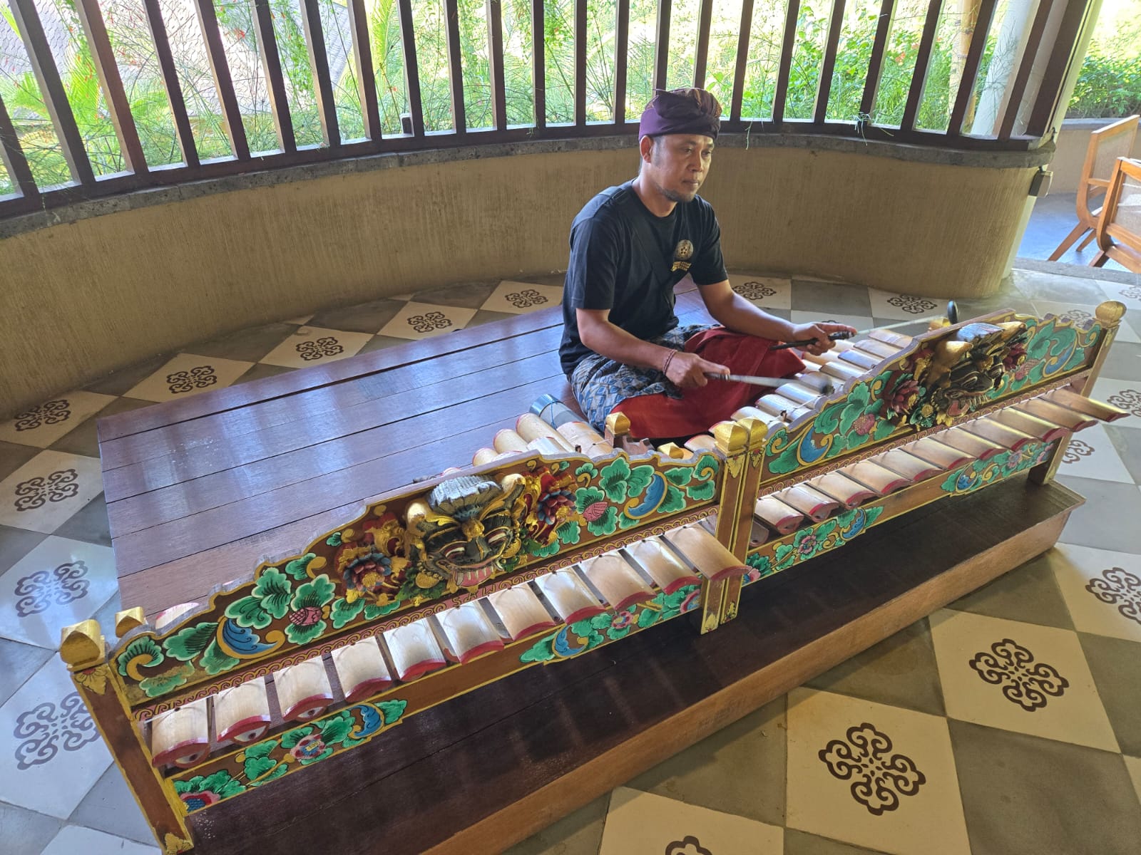 A Balinese musician plays a traditional gamelan instrument indoors, surrounded by intricate carvings and patterned floor tiles. 