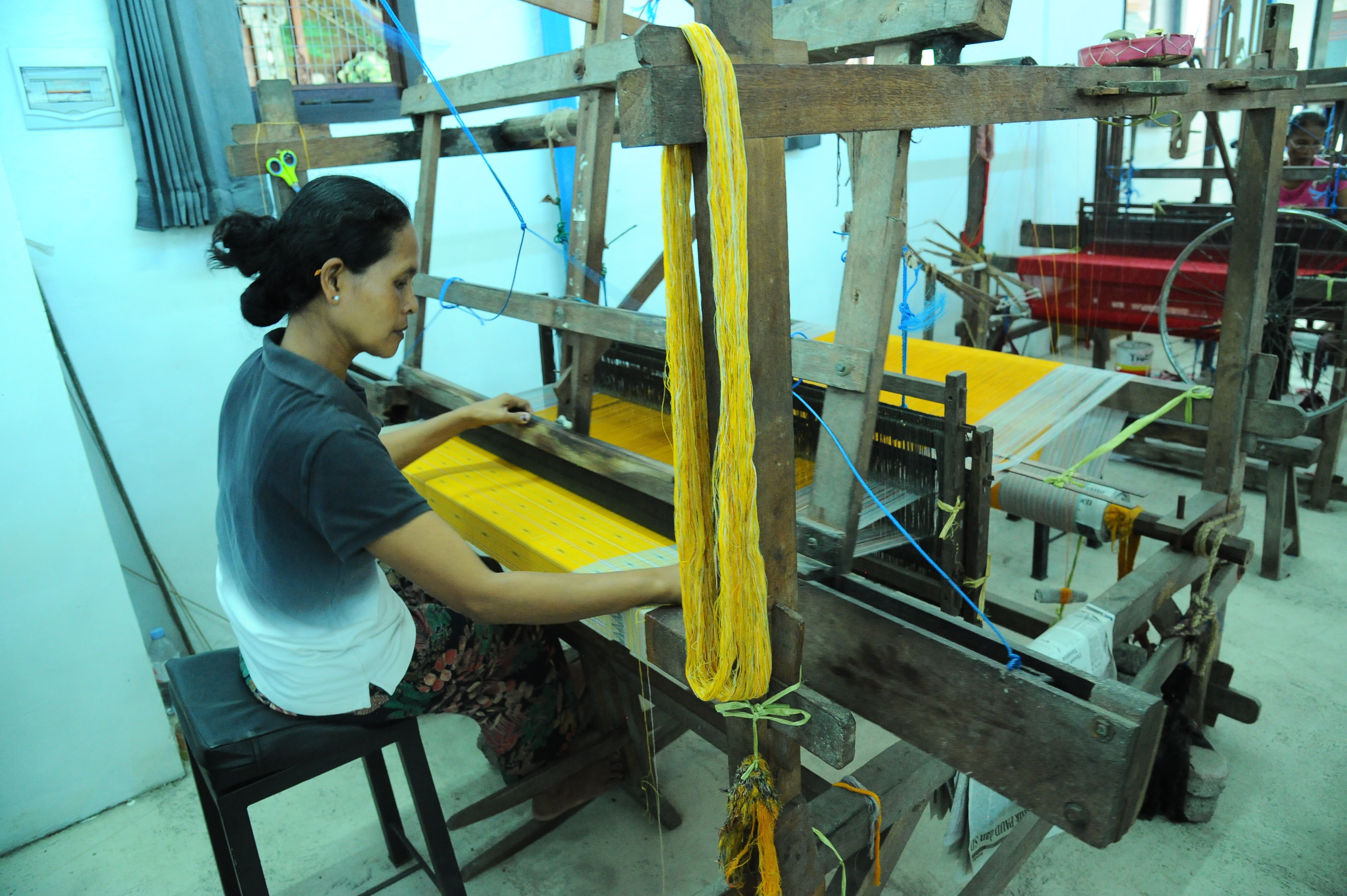 An Indonesian woman carefully weaves bright yellow threads on a traditional wooden loom inside a small textile workshop.