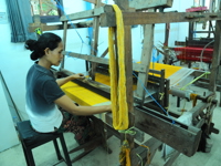 An Indonesian woman carefully weaves bright yellow threads on a traditional wooden loom inside a small textile workshop.