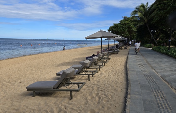 A serene beachfront promenade in Bali with sun loungers, umbrellas, and a cyclist passing under palm trees beside the ocean. 