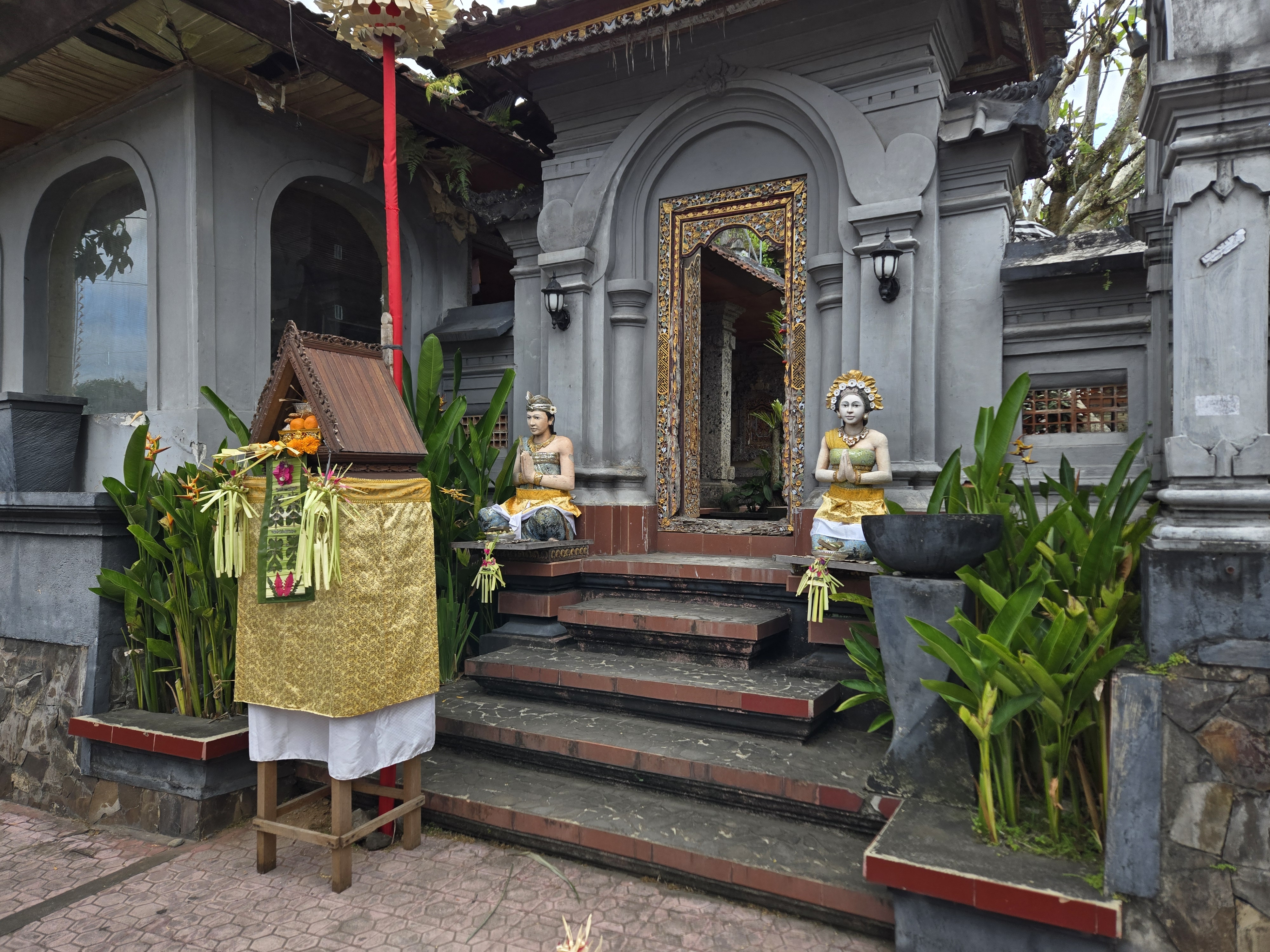 Ornate entrance to a Balinese temple, flanked by statues and an offering shrine, surrounded by tropical greenery. 