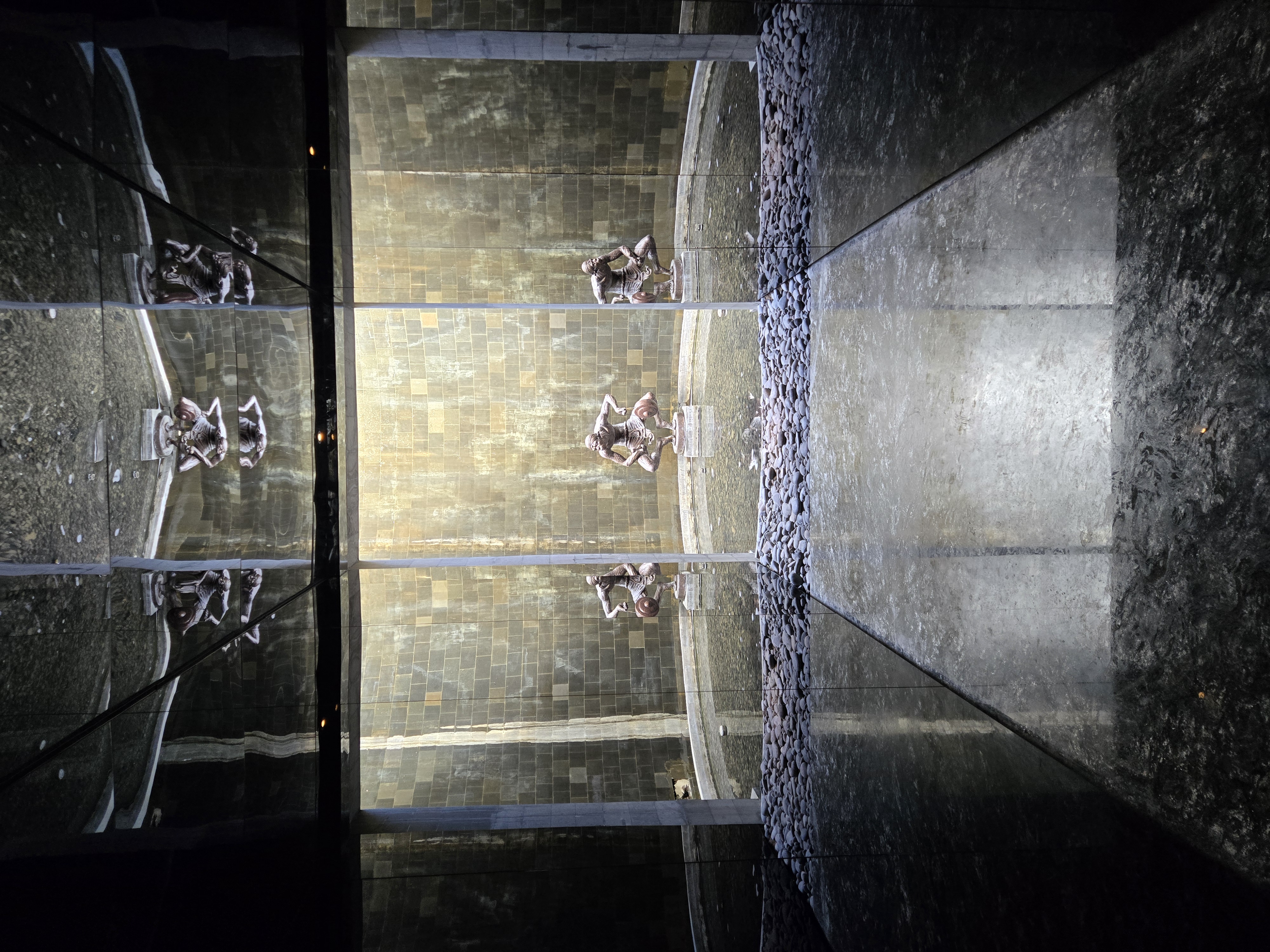 Modern art sculpture reflected in a sleek, water-lined space with stone walls and dramatic lighting, located in Indonesia. 