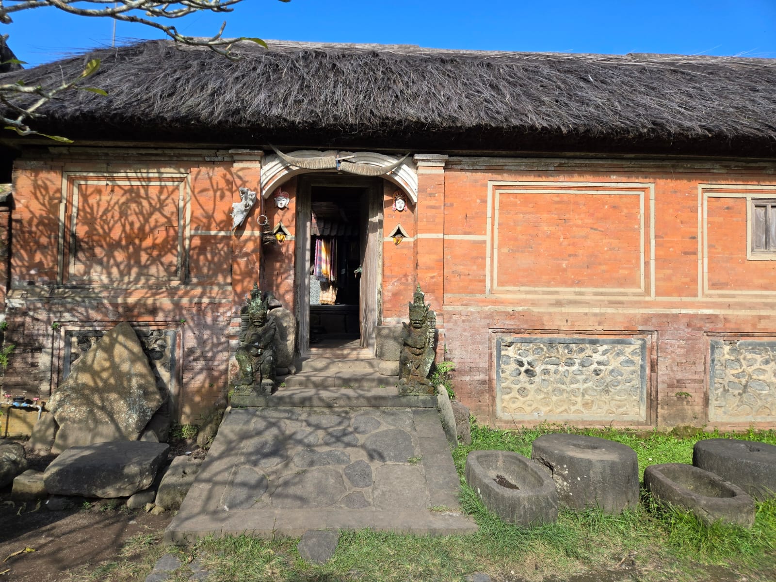 A traditional Balinese home entrance with stone path, red brick walls, and a thatched roof under a clear blue sky in Indonesia.