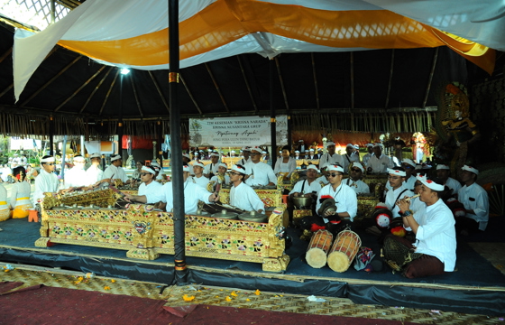 Balinese musicians dressed in white perform gamelan music during a traditional temple ceremony under a decorative pavilion. 
