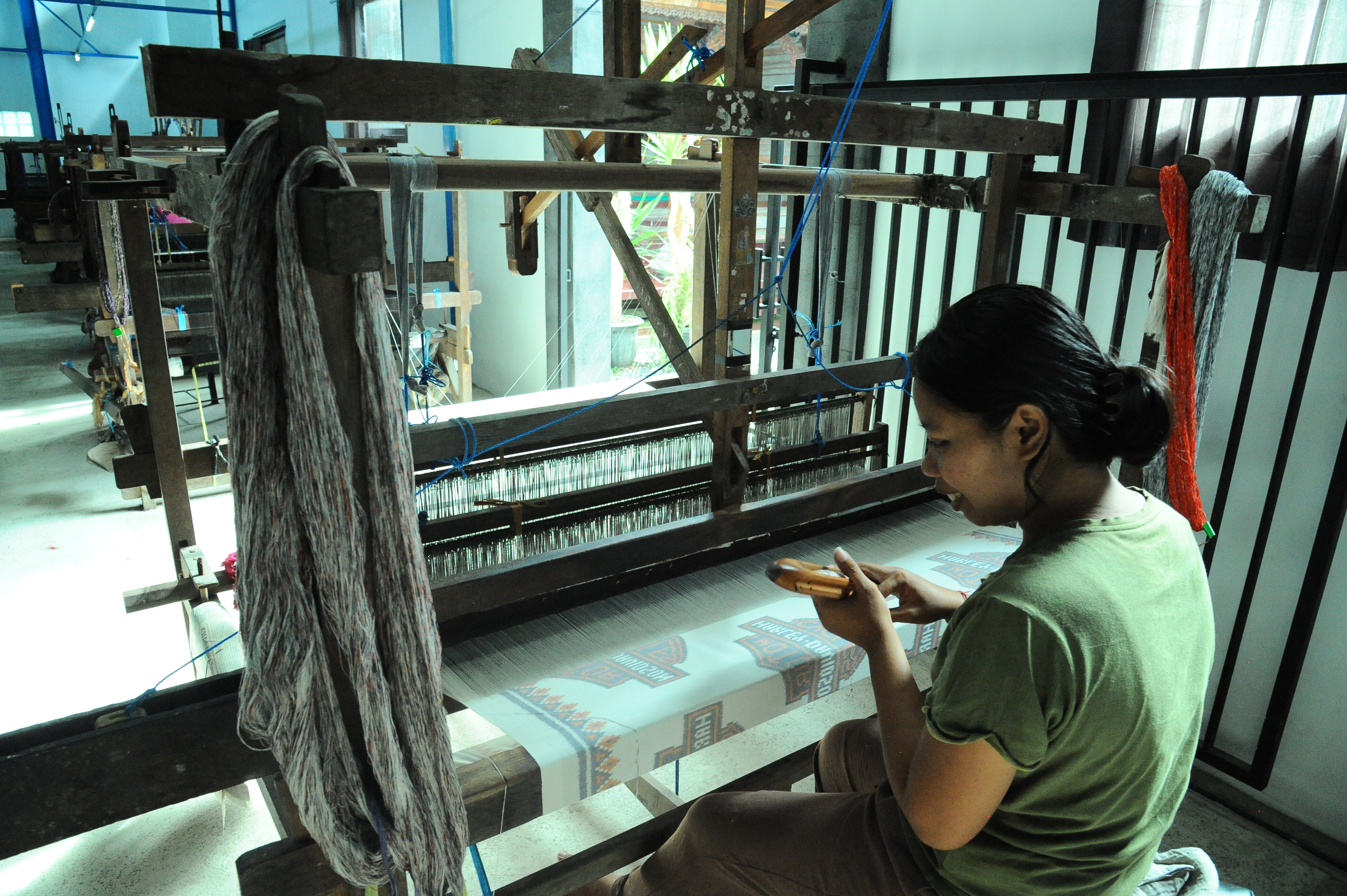 Indonesian woman weaving fabric on a traditional wooden loom in a textile workshop using natural yarns. 