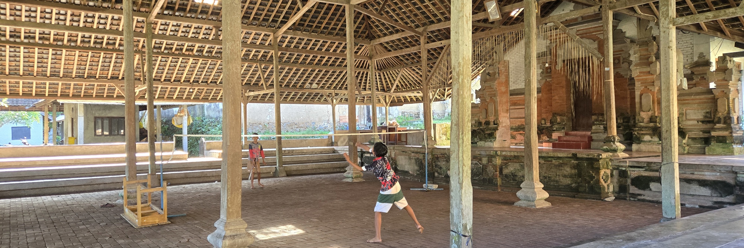 Children playing badminton inside a traditional wooden pavilion with high thatched roof in Bali, Indonesia. 