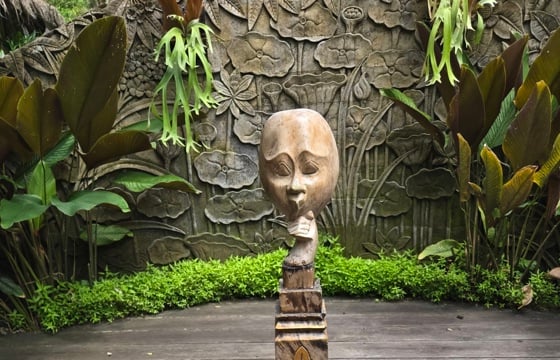A carved wooden sculpture of a face stands on a decorative pedestal, framed by lush tropical plants and a stone floral relief wall. 